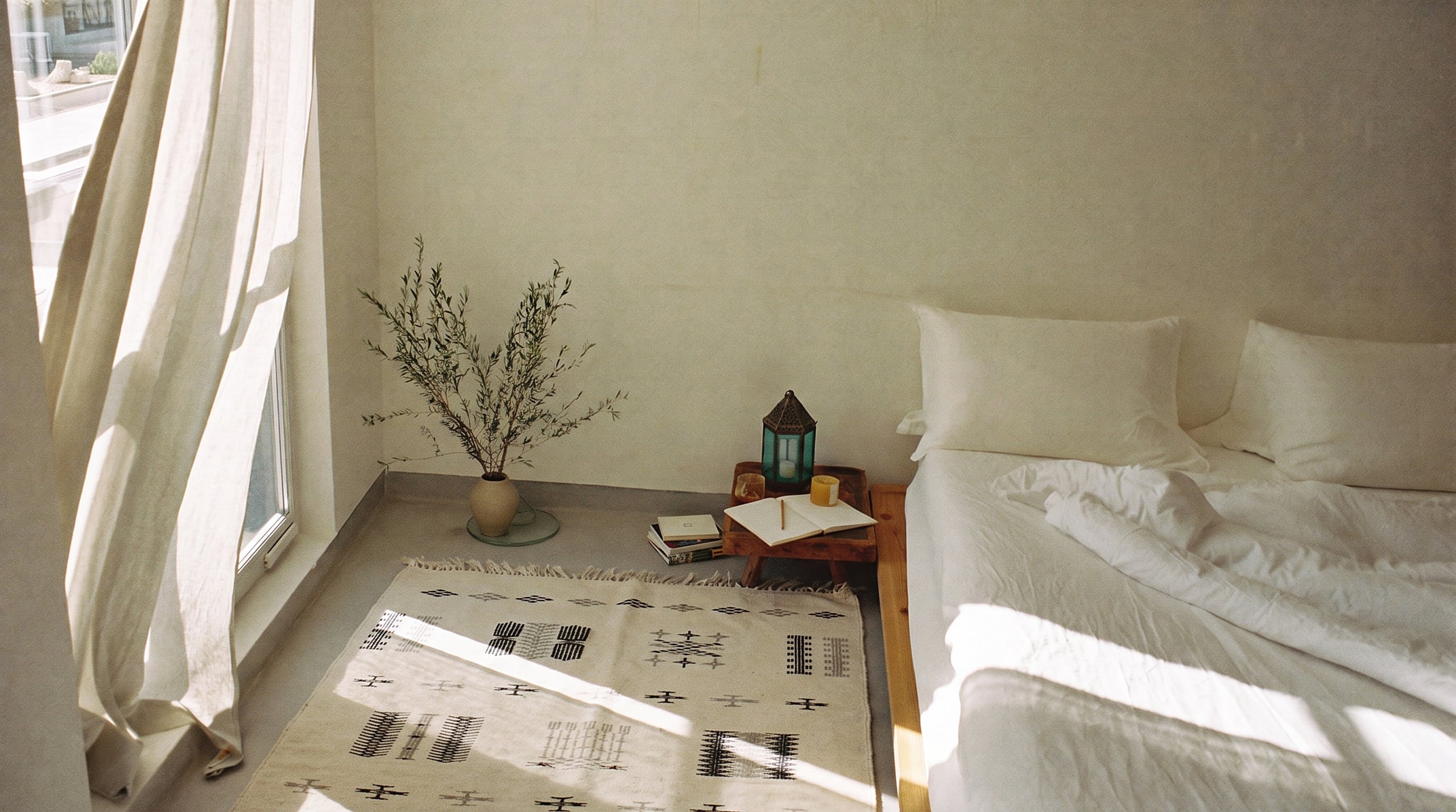 Chambre lumineuse, tapis hanbel cream à motifs noir et gris posé au sol le long d’un lit bas en lin blanc, lanterne et carnet sur un tabouret, grande fenêtre au voilage
